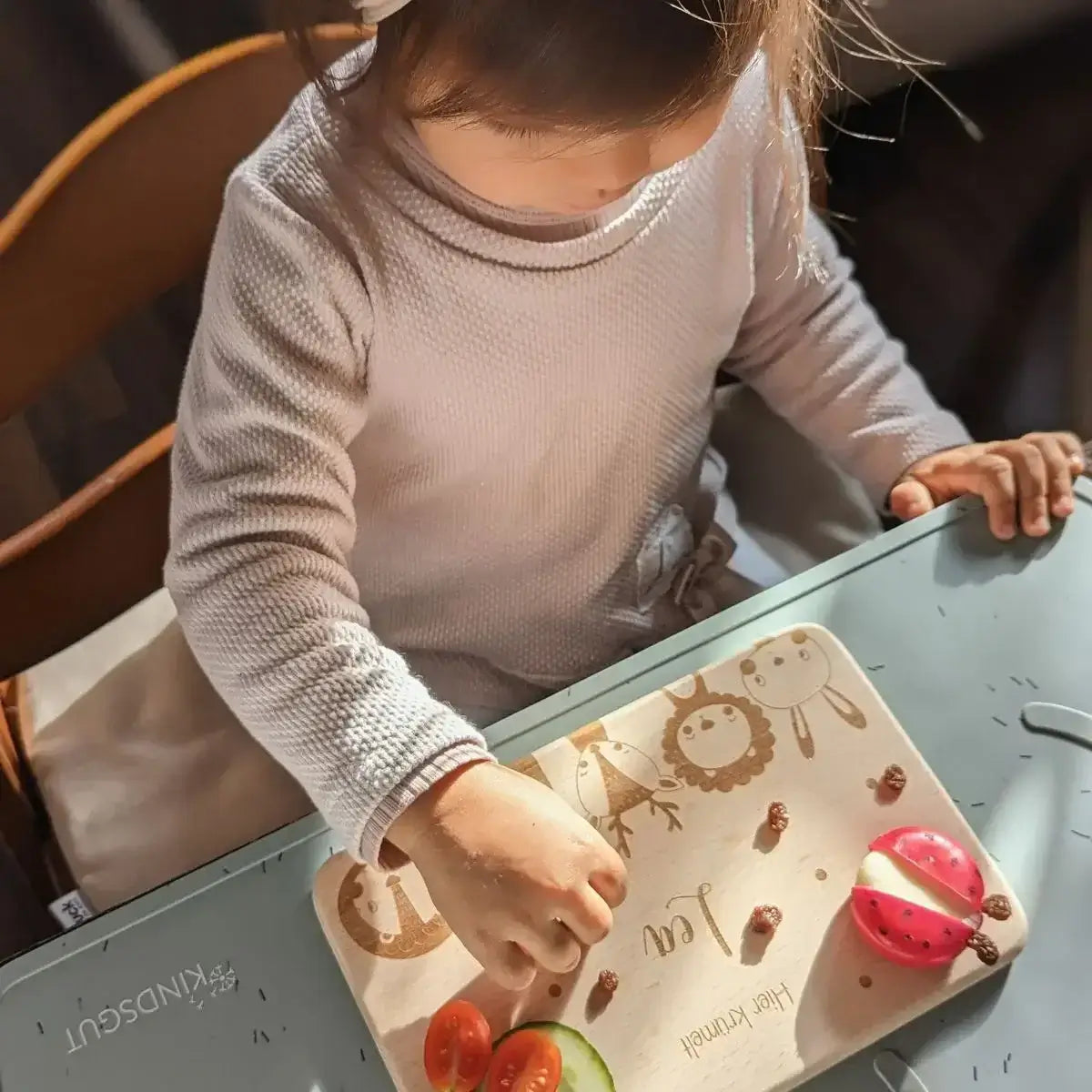 Kleinkind mit personalisiertem Frühstücksbrettchen beim Essen - niedliches Kind in hellem Langarmshirt verwendet ein Holzbrettchen mit Tiermotiven für sein gesundes Frühstück mit Gemüse